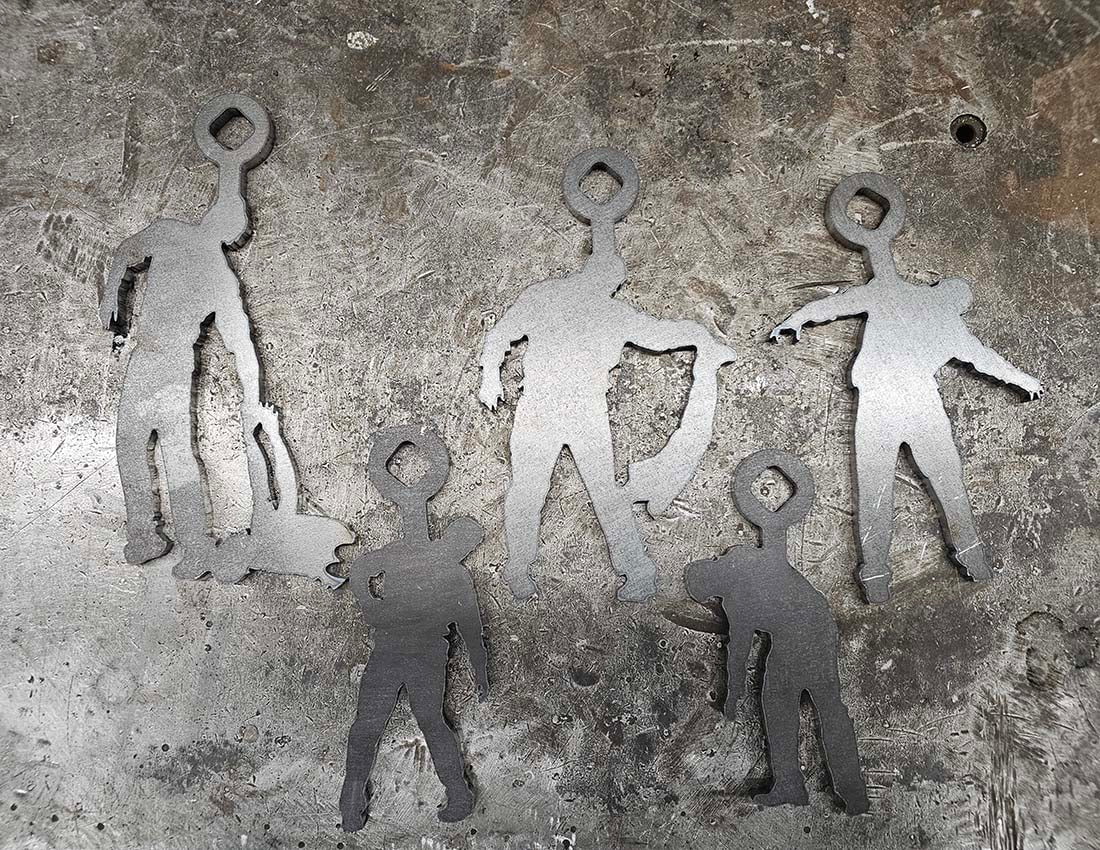 Five cut-out steel zombie targets displayed on a worn metallic surface. Each target has a loop hanger on top, varying in size and pose.