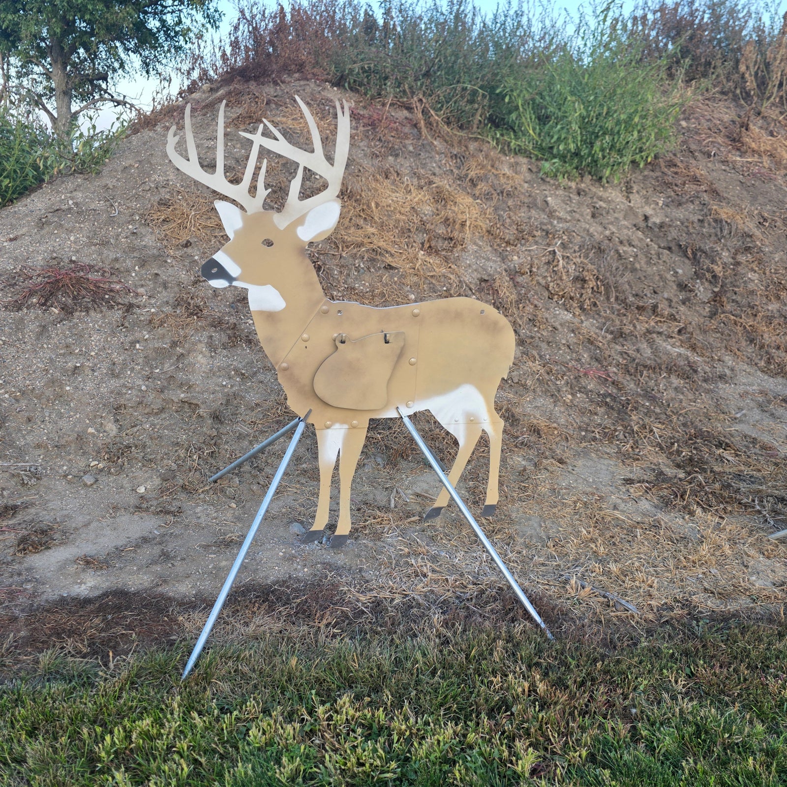A metal deer hunting target shaped like a buck with large antlers, mounted on a tripod stand in an outdoor grassy and dirt setting.